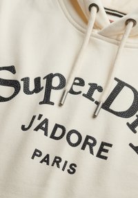 Crème-kleurige hoodie met zwarte, glanzende opdruk "SuperDry J'ADORE PARIS." Voorzien van een trekkoord met metalen punten en een zachte stof textuur.