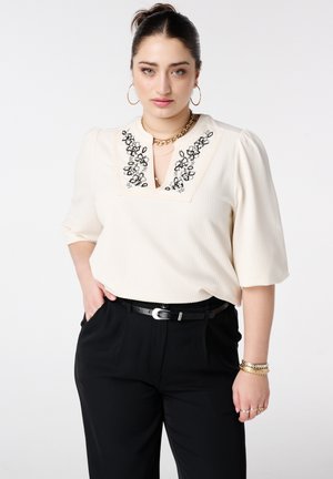 Femme aux cheveux attachés portant un chemisier crème avec une broderie florale noire et un pantalon noir, accessoirisée de bijoux en or.