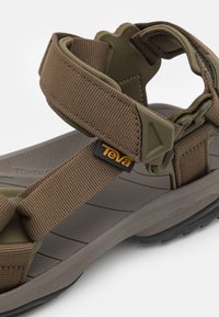 Hnědá sandál se nastavitelními nylonovými popruhy a plastovými detaily. Funkce zahrnuje polstrovanou šedou podrážku s texturovaným povrchem pro lepší grip. Logo Teva je viditelné.