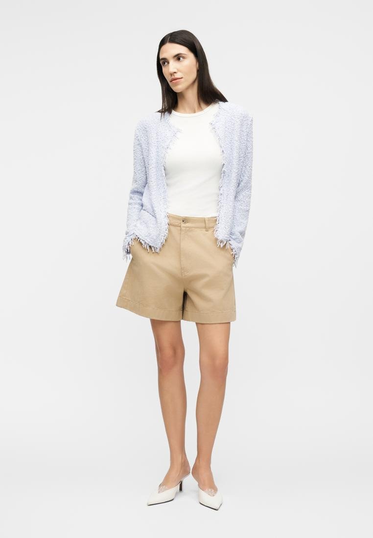 Femme portant un haut blanc, une veste texturée bleu clair, un short beige et des talons blancs pointus, debout les mains dans les poches.