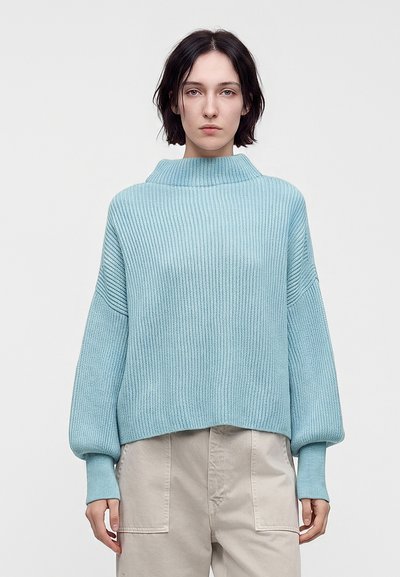Jeune personne aux cheveux courts et foncés, portant un pull bleu clair à côtes et un pantalon beige, tournée vers l’avant sur un fond uni.