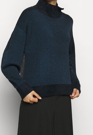 Personne portant un pull à col haut bleu foncé et noir avec des manches longues et des poignets côtelés, assorti à une jupe ou un pantalon noir.