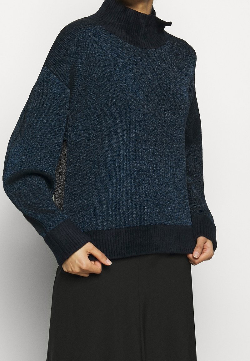 Personne portant un pull à col haut bleu foncé et noir avec des manches longues et des poignets côtelés, assorti à une jupe ou un pantalon noir.