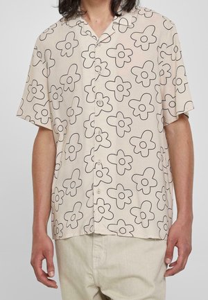 Mann trägt ein beige Hemd mit kurzen Ärmeln und großem abstrakten schwarzen Blumenmuster sowie hellen Hosen.