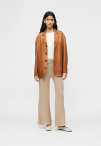 Γυναίκα που στέκεται φορόντας καφέ δερμάτινο σακάκι, λευκό ριμπ μπλουζάκι, μπεζ πλεκτό παντελόνι με κορδόνι και μπεζ παπούτσια χωρίς κορδόνια σε απλό φόντο.