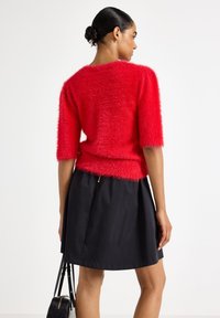 Maglione corto a maniche corte rosso e peloso con una texture morbida, abbinato a una gonna plissettata nera. La modella tiene in mano una borsa nera con motivo coccodrillo.