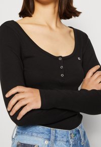 Schwarzes Langarm-Henley-Shirt aus geripptem Material, mit einem Rundhalsausschnitt und drei Metallknöpfen auf der Vorderseite.