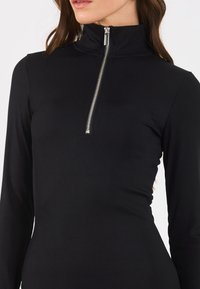 Top negru cu mânecă lungă și texturat, având un guler înalt și un fermoar argintiu la gât, oferind un design ajustat.