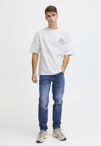 Hvid bomulds T-shirt med rund halsudskæring og korte ærmer, med et grønt og blåt hjerte-logo. Kombineret med blå denim jeans og beige sneakers.