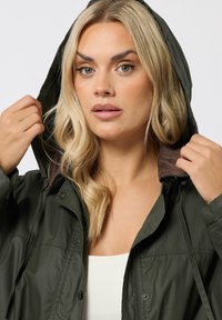 Veste à capuche vert olive avec fermeture éclair à l'avant et boutons-pression. Doublure intérieure marron, texture légère et cordon de serrage ajustable au niveau de la capuche.