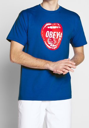 Camiseta azul con un gráfico rojo de una boca abierta y el texto blanco "OBEY" llevada por una persona con los brazos cruzados y pantalones blancos.