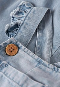 Svetlo modre jeans kombinezoni imajo naguban trak in zapirajoč gumb iz lesa, ki prikazujejo gladko teksturo in klasične oblikovne elemente.
