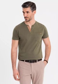 Camiseta de manga corta verde oliva con escote redondeado, tapeta de tres botones en un tono más claro y un bolsillo en el pecho. Llevada con pantalones beiges.