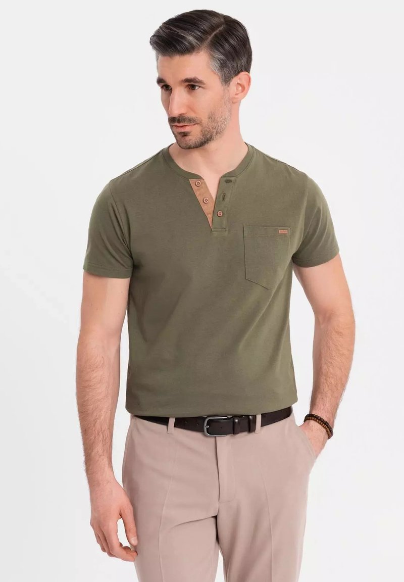 Camiseta de manga corta verde oliva con escote redondeado, tapeta de tres botones en un tono más claro y un bolsillo en el pecho. Llevada con pantalones beiges.