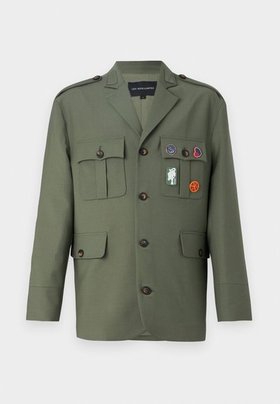 Veste vert olive avec quatre poches avant, fermeture à boutons et patchs décoratifs de différentes couleurs. Elle comporte des épaulettes aux épaules.