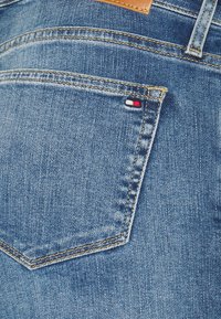 Modré džínové kalhoty s viditelnou zadní kapsou, s kontrastním béžovým prošíváním a malou červenou, bílou a modrou štítkovou značkou na kapse.