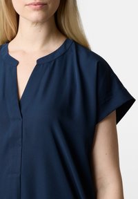 Navyblaue Bluse mit V-Ausschnitt, kurzen Kapuzenärmeln und glattem Stoff. Mit feinen Nähdetails entlang der Knopfleiste.