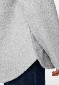 Pull gris fluffy avec une surface texturée et douce, présentant une coupe décontractée et un ourlet arrondi. Associé à un jean en denim foncé.