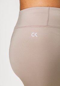 Legging beige clair avec une taille haute, texture lisse et logo "CK" blanc à l'arrière. Les coutures apportent de la structure.