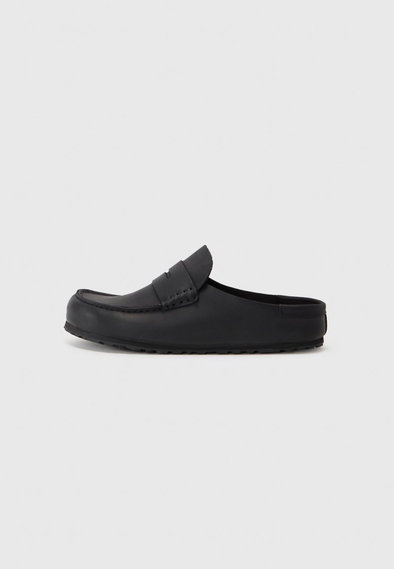 Chaussure noire en cuir à enfiler avec un bout rond, des coutures décoratives et une semelle en caoutchouc texturée. Surface lisse avec des accents minimaux.