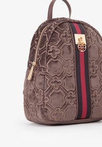 Zaino in suede color taupe con texture decorata e dettagli in oro. Presenta una striscia centrale rossa e nera, chiusura con zip e spallacci imbottiti.