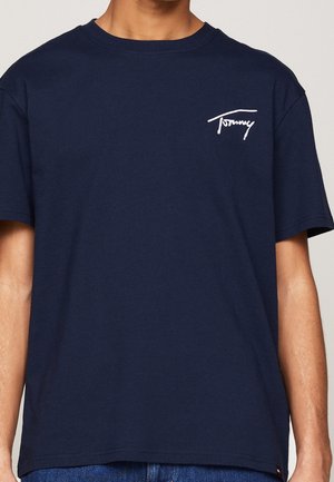 Mężczyzna w jednolitej granatowej koszulce z małym białym logo "Tommy" na górnej lewej części klatki piersiowej i niebieskich dżinsach.