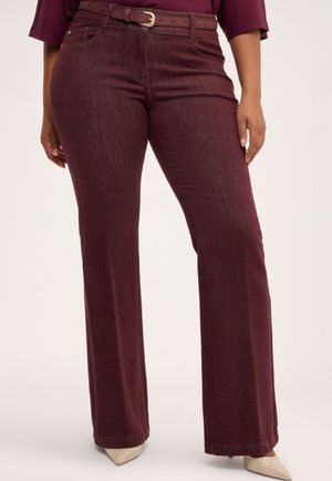 Femme portant un pantalon large taille haute bordeaux avec une ceinture assortie et des talons pointus crème, debout devant un fond uni.