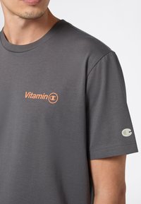 Mężczyzna w ciemnoszarej koszulce z logo "Vitamin C" w pomarańczowym kolorze na piersi oraz małym logo Champion na lewej rękawie.