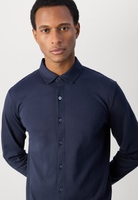 Polo a maniche lunghe blu navy con colletto, chiusura a bottoni e trama liscia. Ha una vestibilità aderente al corpo, che mette in mostra un design minimalista.