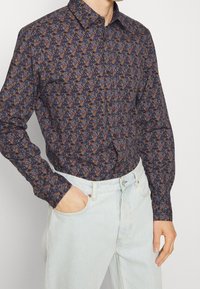 Chemise à boutons dans un tissu bleu foncé avec un motif floral coloré. Présente des manches longues, un col, et une coupe ajustée. Portée avec un jean clair.