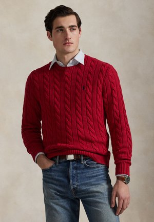 Homme portant un pull en maille torsadée rouge par-dessus une chemise à rayures avec col, jean bleu et montre-bracelet, debout avec une main dans la poche.