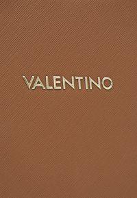 Smeša smeđeg teksturiranog kože s dijagonalnim linijama; sadrži zlatni logotip "VALENTINO" u modernom tipu pisma, centriran i istaknut.