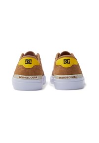 Brune ruskindssneakers med hvide gummisåler og gule hældetaljer med et sort logo. Bagfra vises teksturerede detaljer og branding.