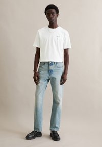 T-shirt bianca con scollo rotondo e piccolo logo, abbinata a jeans denim azzurri. Il modello indossa scarpe slip-on nere con suola testurizzata.