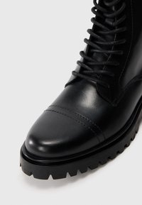 Bota de cuero negro con punta redonda, superficie texturizada y seis ojales de metal. Suela de goma con un dibujo pronunciado para mayor tracción.