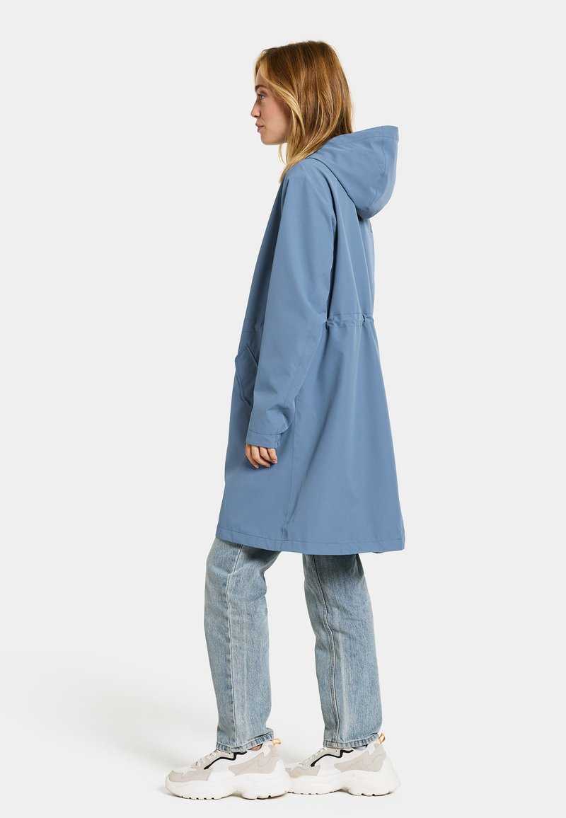 Hellblauer Regenmantel mit Kapuze, Seitentaschen und lockerer Passform, kombiniert mit hellblauen Jeans und sneakers in neutralen Farbtönen.