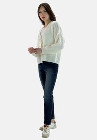 Flauschige gestrickte cremefarbene Strickjacke mit rundem Halsausschnitt und Bindegurt, kombiniert mit dunkelblauen Slim Jeans und hellen Sneakers.