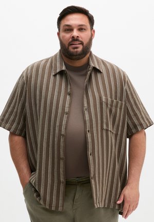 Mężczyzna z brodą, ubrany w brązową prążkowaną koszulę z krótkim rękawem na brązowy t-shirt i zielone spodnie, stoi z rękami w kieszeniach.