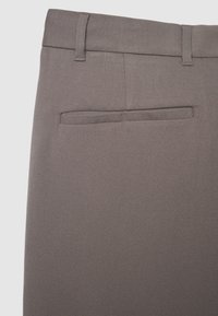 Nahaufnahme von braunen Stoffhosen, die den hinteren Bund, die Gürtelschlaufen und das Detail der einzelnen Paspeltasche zeigen.