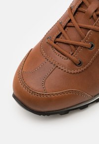 Ruskea nahkakenkä, jossa on teksturoidun pinnan, pyöreä kärki ja monimutkainen ompelu. Kenkässä on ruskeat nauhat ja musta kumipohja.