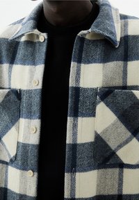 Giacca camicia in lana a quadri nei colori crema, blu navy e azzurro. Presenta un colletto, chiusura con bottoni e due tasche frontali. Tessuto con texture.