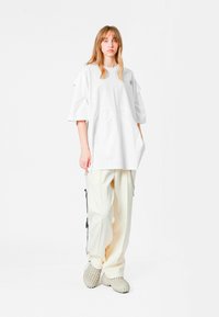 Camiseta blanca de gran tamaño con hombros caídos y detalles recortados, combinada con pantalones anchos de color crema y calzado texturizado gris.