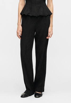 Žena v voľných čiernych nohaviciach, čiernych špicatých balerínach a čiernom tielku bez rukávov s vlnkovaným lemom stojaca pred jednofarebným pozadím.