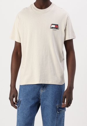 Beige Baumwoll-T-Shirt mit Rundhalsausschnitt und kurzen Ärmeln. Es hat einen bedruckten Grafik in Schwarz, Rot und Weiß auf der linken Brust.