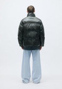 Fodrad svart pufferjacka med hög krage, med en blank yta, i kombination med vida ljusblå jeans. Bakre vy visas.