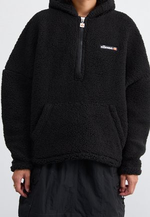 Osoba nosząca czarną polarową bluzę z kapturem, z kieszenią z przodu i suwakiem do połowy długości, z logo Ellesse na piersi.