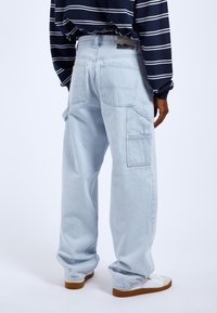 Pantalones cargo de mezclilla azul claro con corte relajado, múltiples bolsillos y una cinturilla con la marca "DIE DILIG." Llevados con zapatillas blancas.