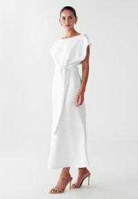Bijela haljina s drapiranim kragnom, vezanim strukom i lepršavom suknjom. Nosi se uz bež sandale na petu. Glatka tekstura tkanine.
