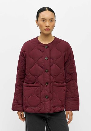 Femme portant une veste matelassée rouge foncé avec de gros boutons et des poches avant, se tenant devant un fond clair uni.