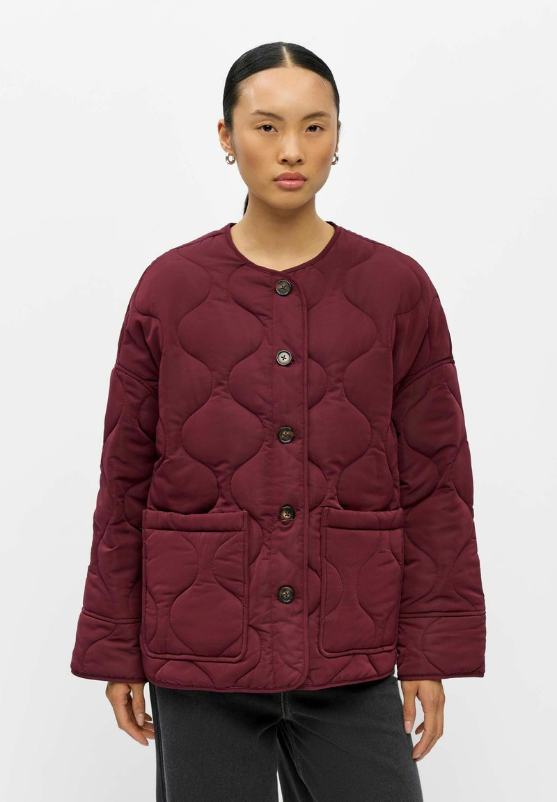 Femme portant une veste matelassée rouge foncé avec de gros boutons et des poches avant, se tenant devant un fond clair uni.